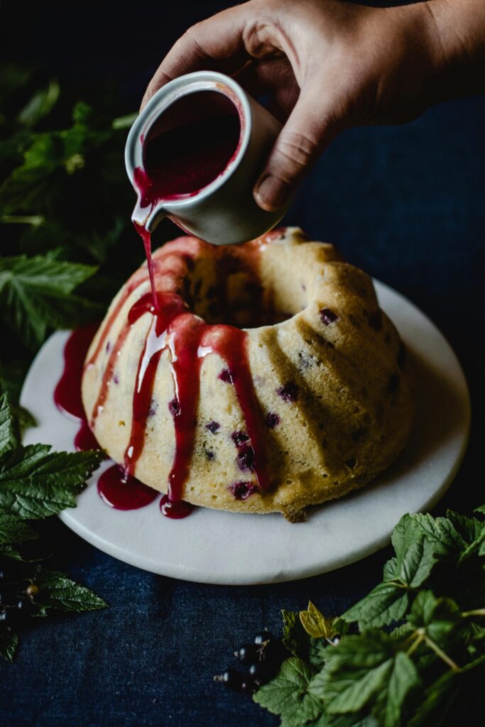 Hand pouring berry sauce over a bundt cake on a marble plate surrounded by fresh green leaves