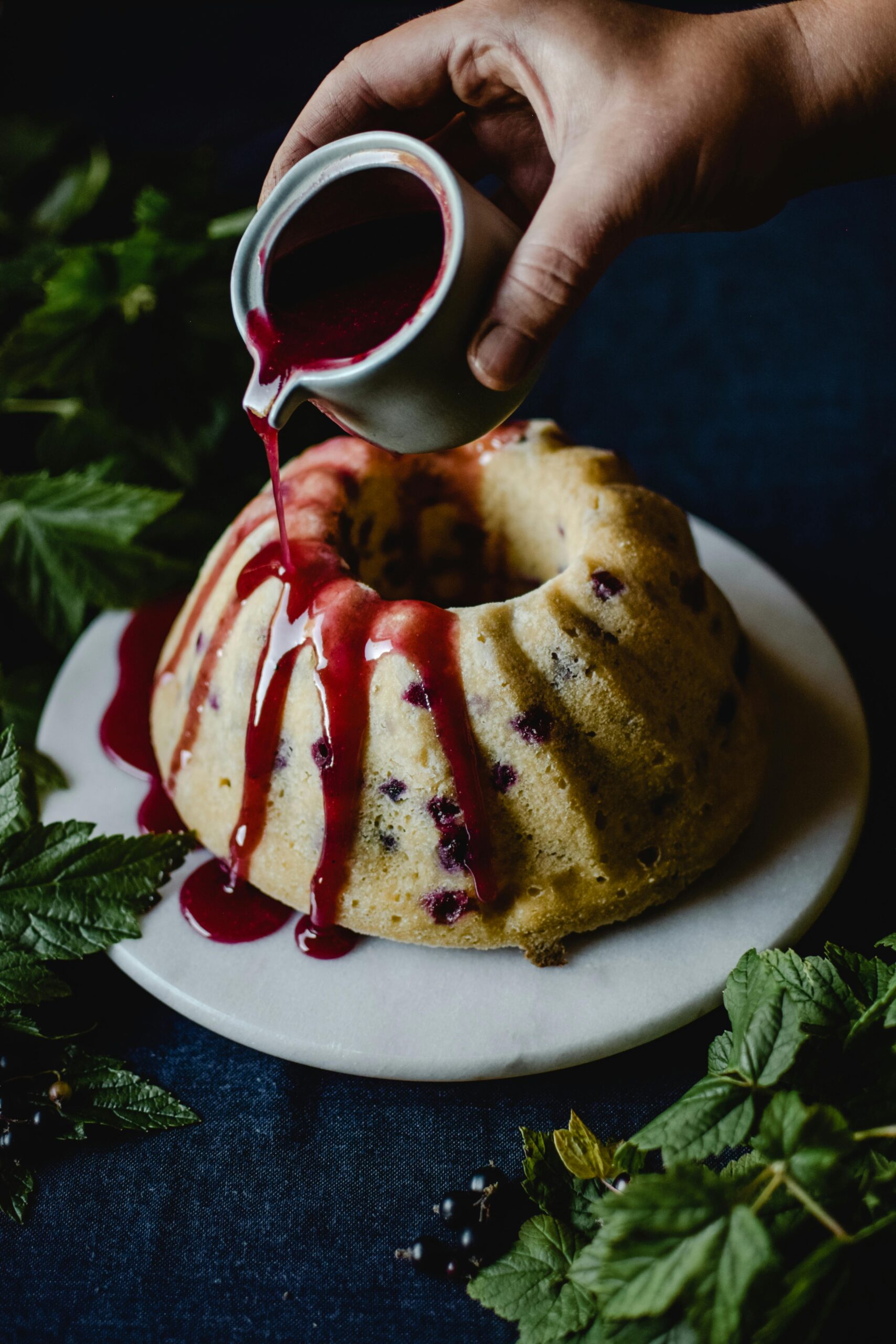 Hand pouring berry sauce over a bundt cake on a marble plate surrounded by fresh green leaves