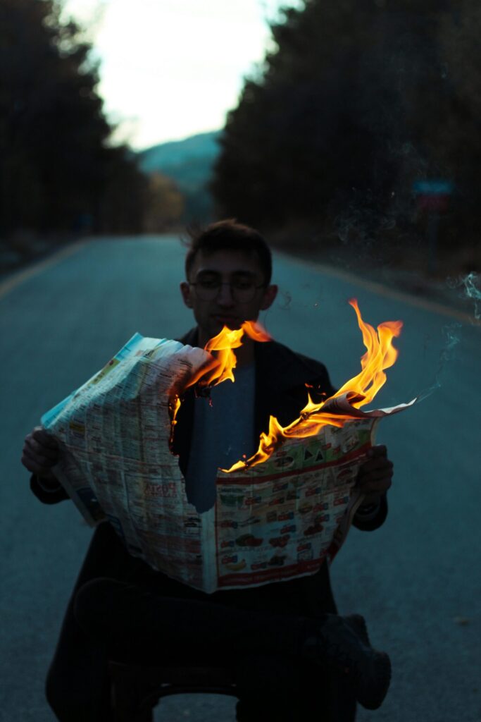 Man in glasses reading a newspaper engulfed in flames while standing on an empty country road
