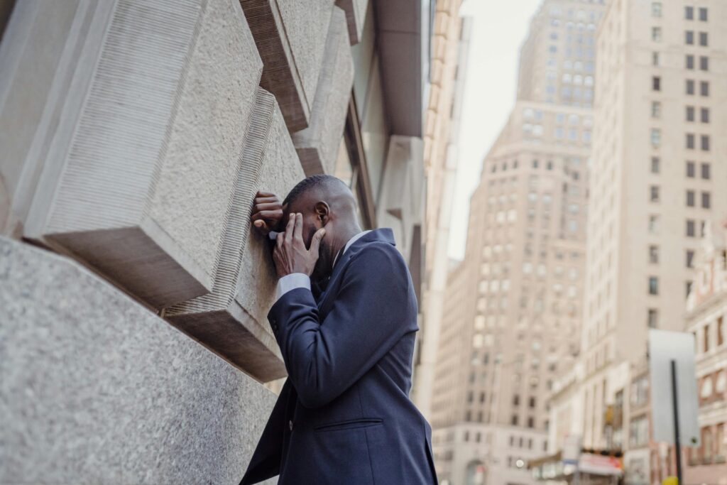 Businessman in a navy suit resting his head against a stone wall amid city skyscrapers
