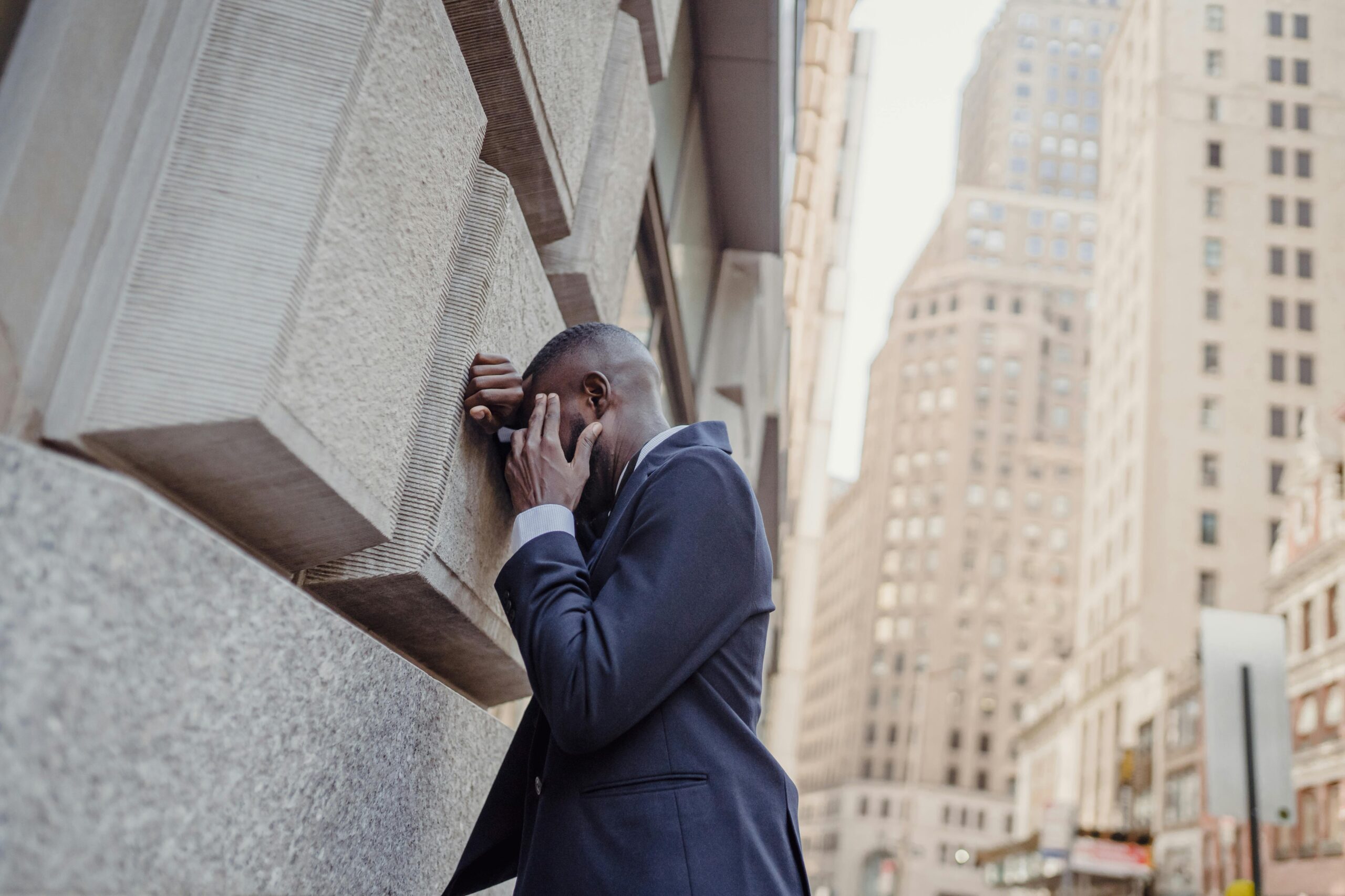 Businessman in a navy suit resting his head against a stone wall amid city skyscrapers