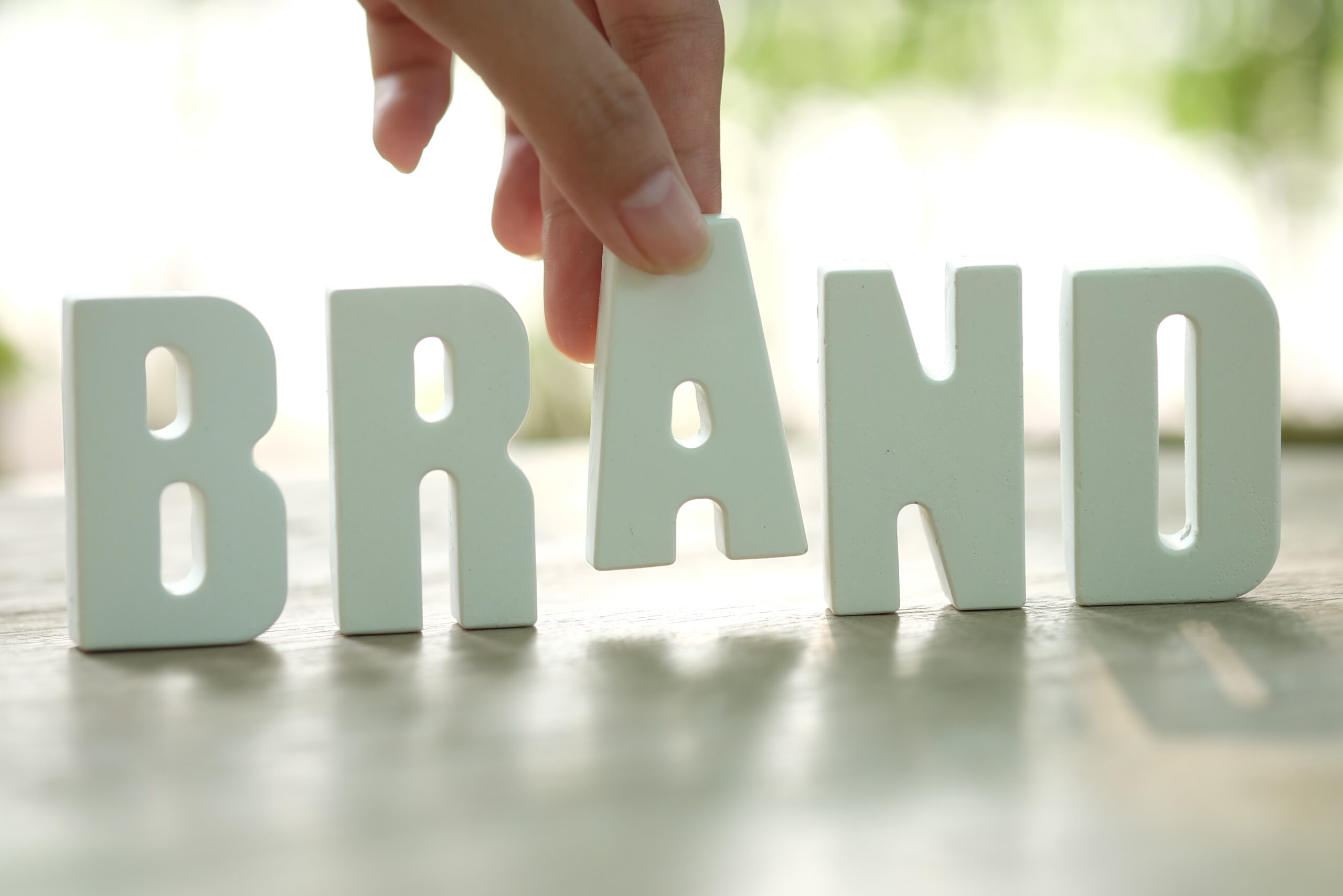 Hand placing wooden block letters spelling BRAND on a wooden table with soft green background